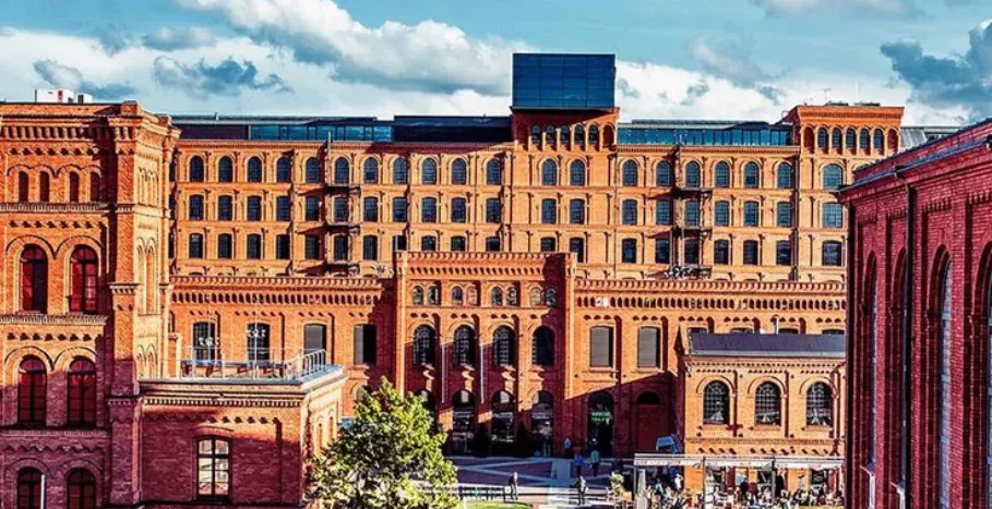 Large complex of red brick industrial buildings under a blue sky with white clouds. People gather in the foreground.