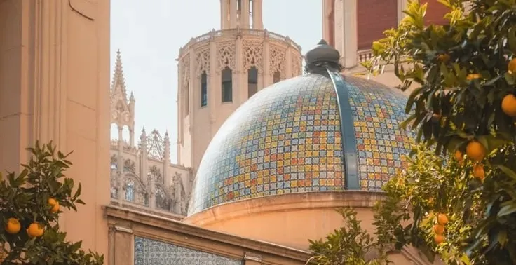 Ceramic tiled dome of the Mercado Central in Valencia framed by ripe orange trees and towers.