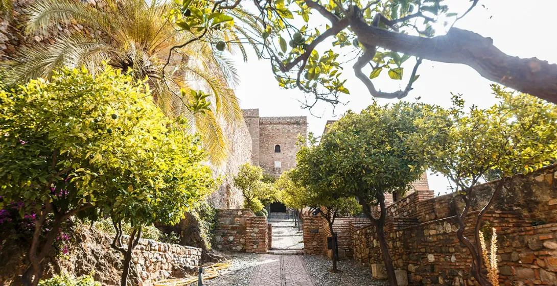 Cobblestone path leading to an old stone fortress in Malaga, flanked by citrus and palm trees and brick walls.