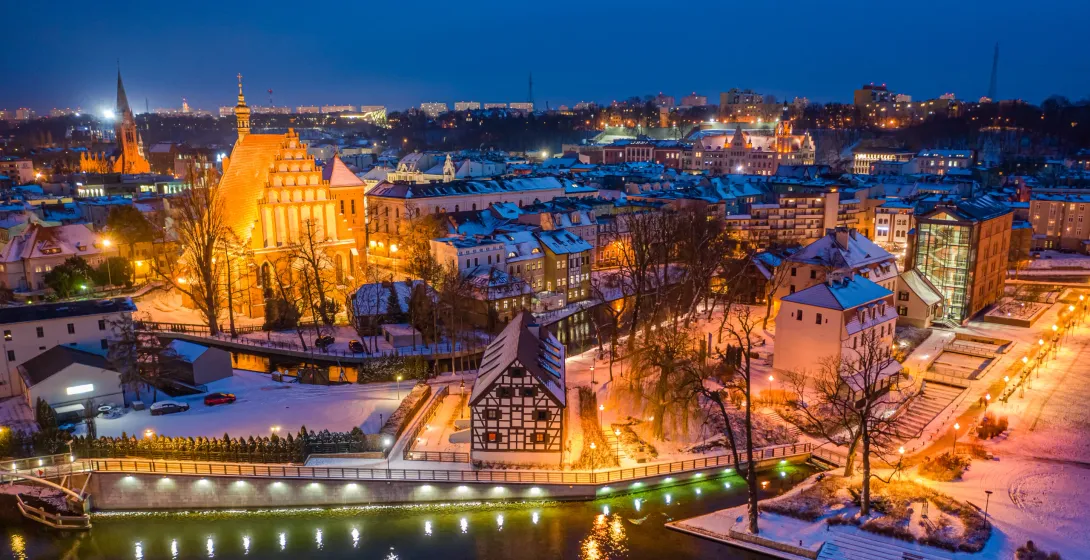 Aerial view of snowy Bydgoszcz Old Town at dusk, with illuminated historic buildings and a river reflecting lights.