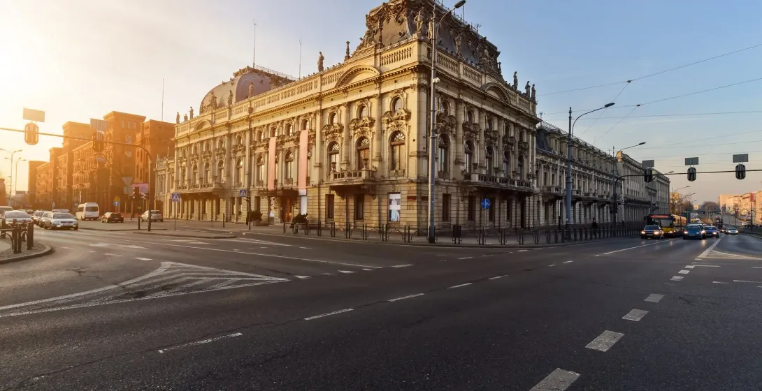 Ornate, light-colored neo-renaissance palace on a sunny city street corner with traffic lights and moving cars.