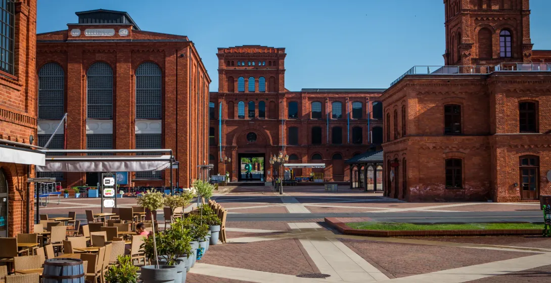 Red brick industrial architecture and a paved square with outdoor cafe seating under a clear blue sky in Lodz.