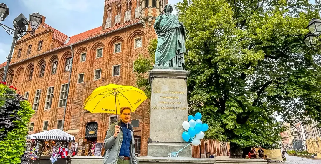 Statue of Nicolaus Copernicus in front of the brick Gothic Old Town Hall in Toruń, with a person holding a yellow umbrella.