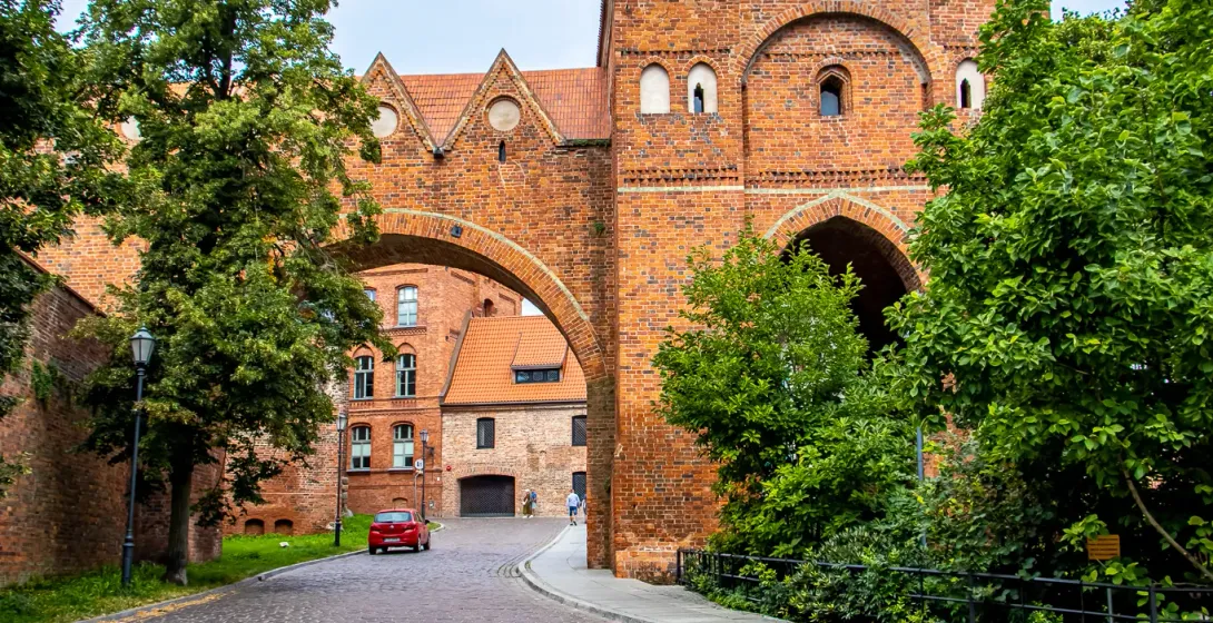 Cobblestone street leads to a historic brick archway gate with red tiled roof, flanked by green trees, a yellow umbrella in f