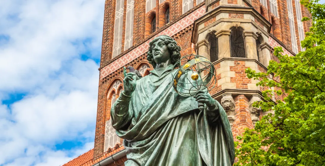 Bronze statue of Nicolaus Copernicus holding an armillary sphere, with a historic brick church tower and blue sky with clouds