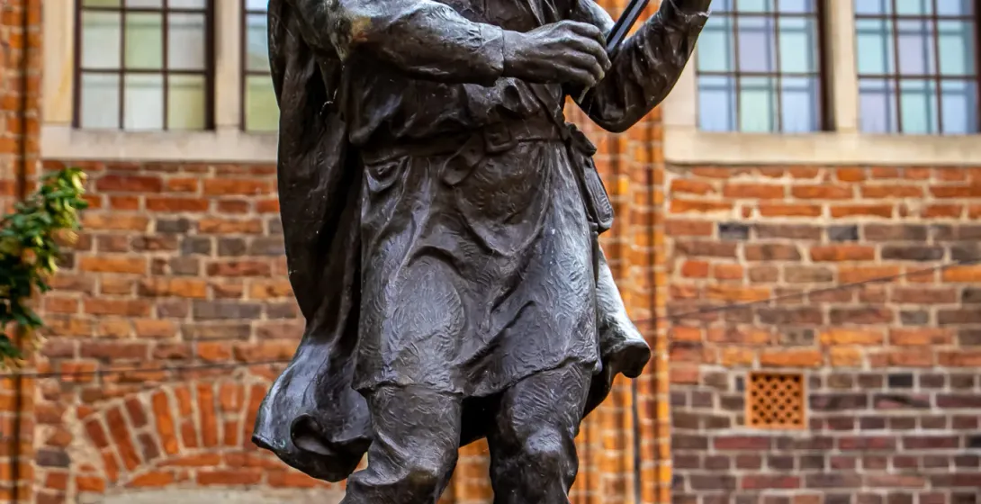 Bronze statue of a barefoot fiddler playing violin, standing on a white pedestal in front of an old brick building with windo