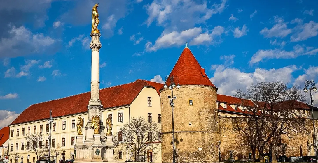 Marian Column with golden statue under a blue sky, next to an old building with a red conical roof in a city square.