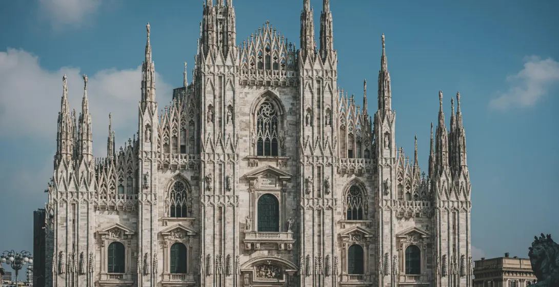 Detailed front facade of Milan Cathedral with intricate Gothic spires, statues, and windows against a blue sky.