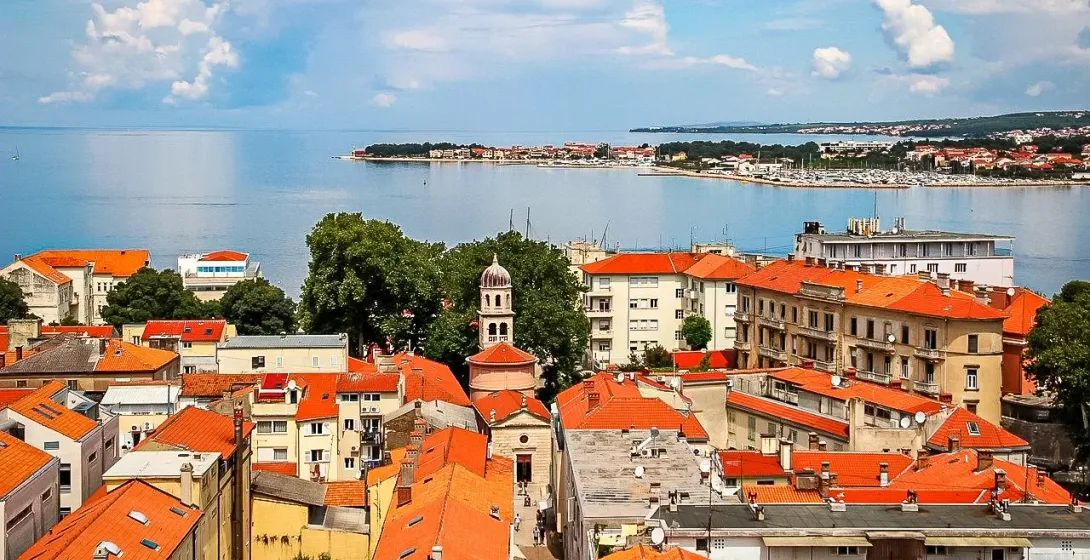 Aerial view of Zadar Old Town with orange tiled roofs, a domed bell tower, and the Adriatic Sea in the background.