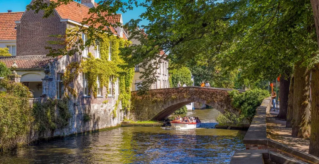 A tour boat with passengers on a canal in Bruges, passing under a stone arch bridge next to an ivy-covered building.