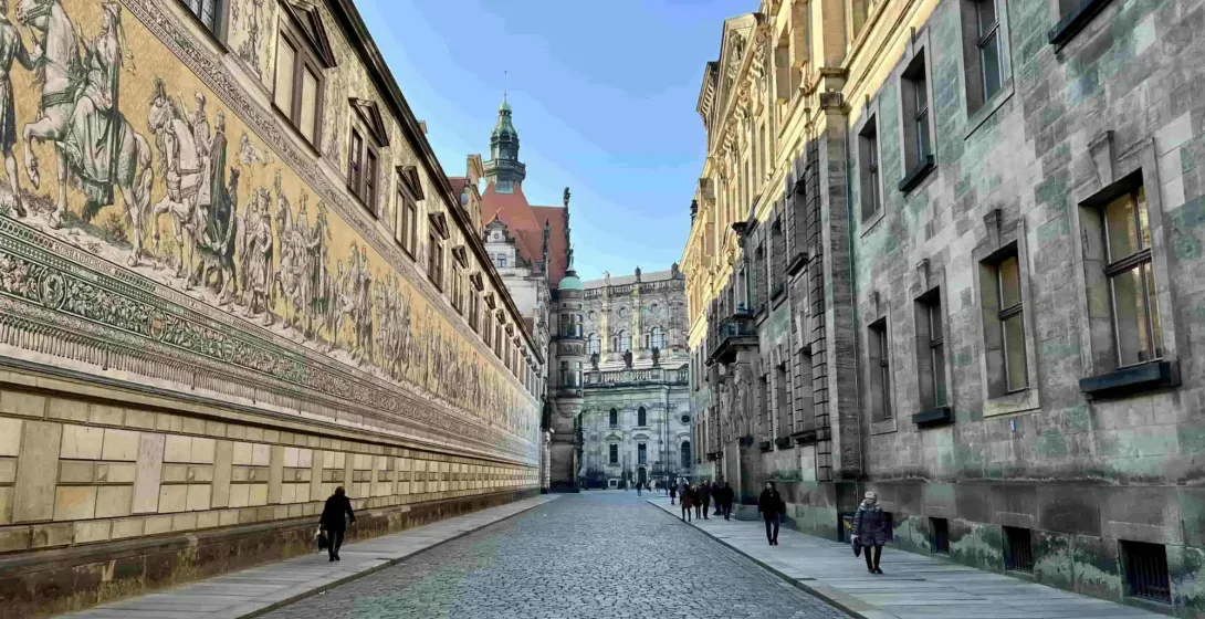 Long, ornate mural depicting a procession of historical figures on a building facade, with a cobblestone street and old build