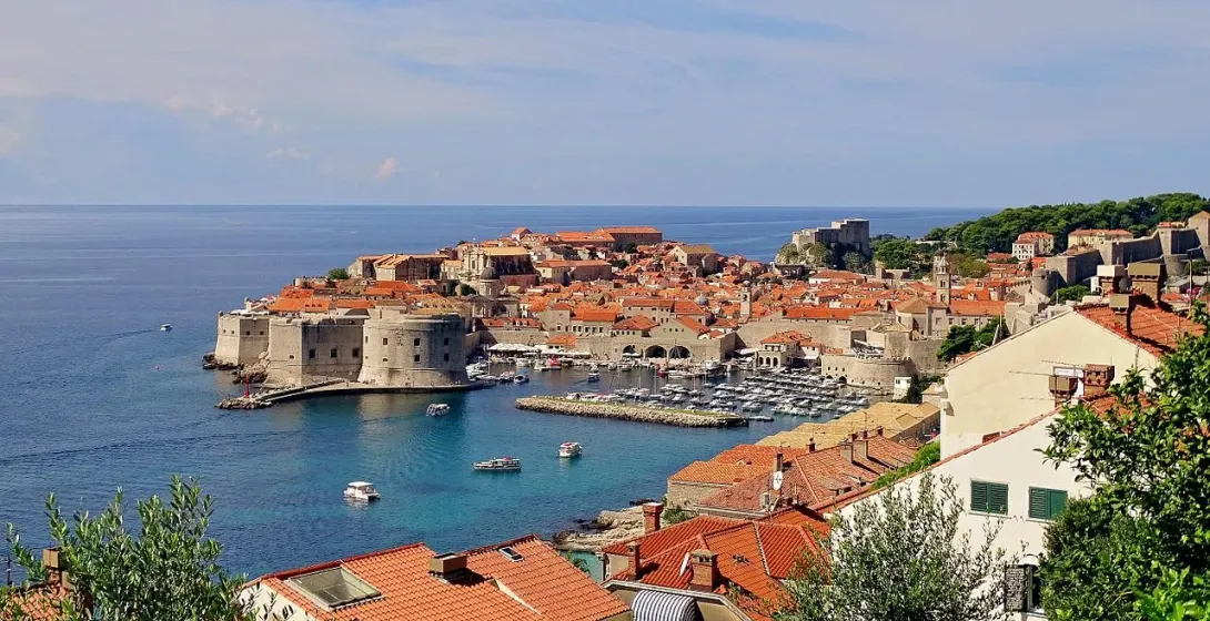 Elevated view of Dubrovnik's Old Town, featuring red-tiled roofs, medieval city walls, a harbor with boats, and the Adriatic