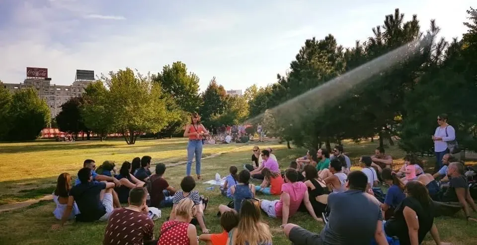 Group of tourists sitting on grass in a park, listening to a guide. A building with a Coca-Cola sign is in the background.