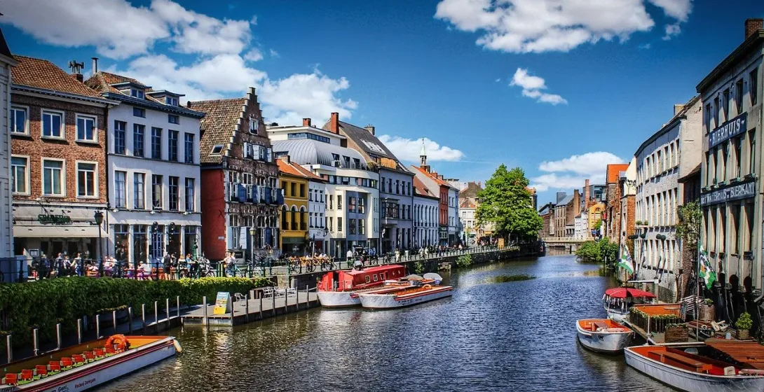 Historic buildings with gabled roofs line a sunny canal with several tour boats and people walking on the banks.