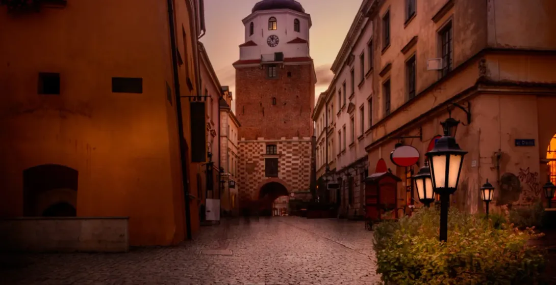 Cobblestone street leading to the Krakow Gate in Lublin Old Town at dusk, flanked by historic buildings and illuminated stree