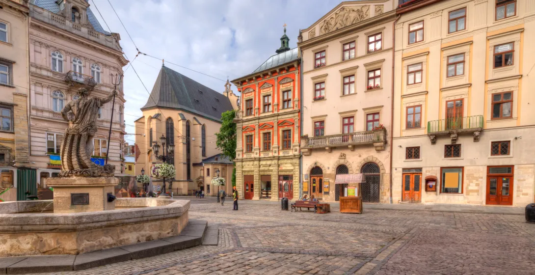 Cobblestone square with a Neptune fountain, surrounded by historic multicolored buildings and a large church under a cloudy s