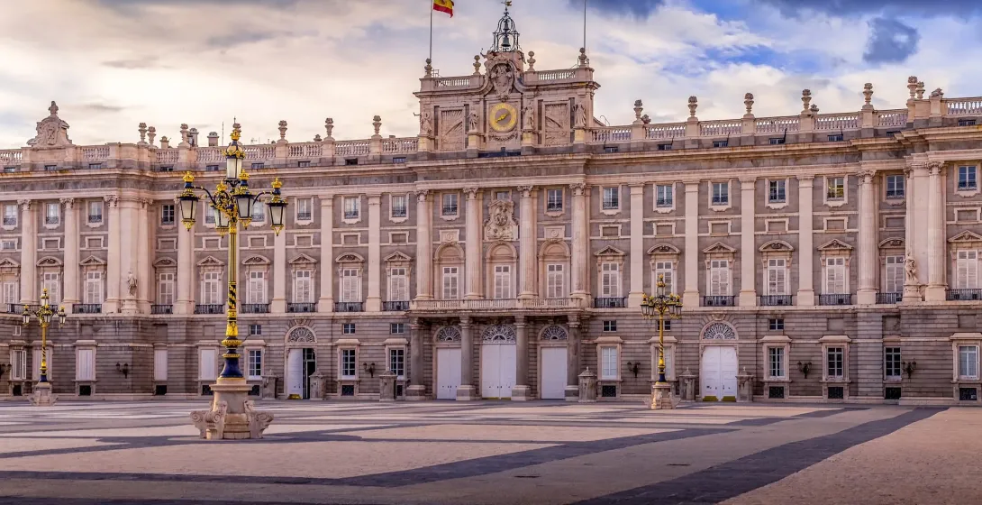 Grand Royal Palace of Madrid with a Spanish flag on top, two ornate street lamps, and a wide empty plaza under a dramatic clo