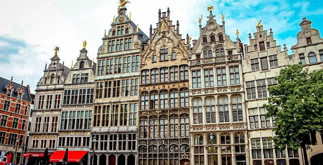 Elaborate historic guildhalls with stepped gables and golden statues line a cobblestone market square in Antwerp. People walk