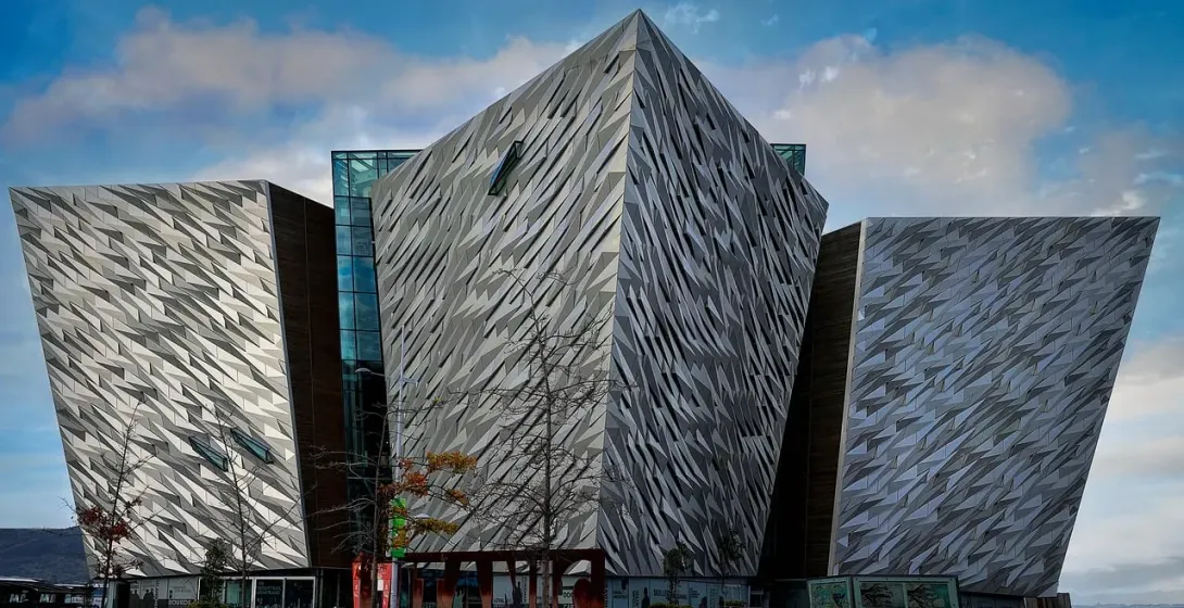 The Titanic Belfast building with its angular, faceted silver panels under a partly cloudy sky, with bare trees in the foregr