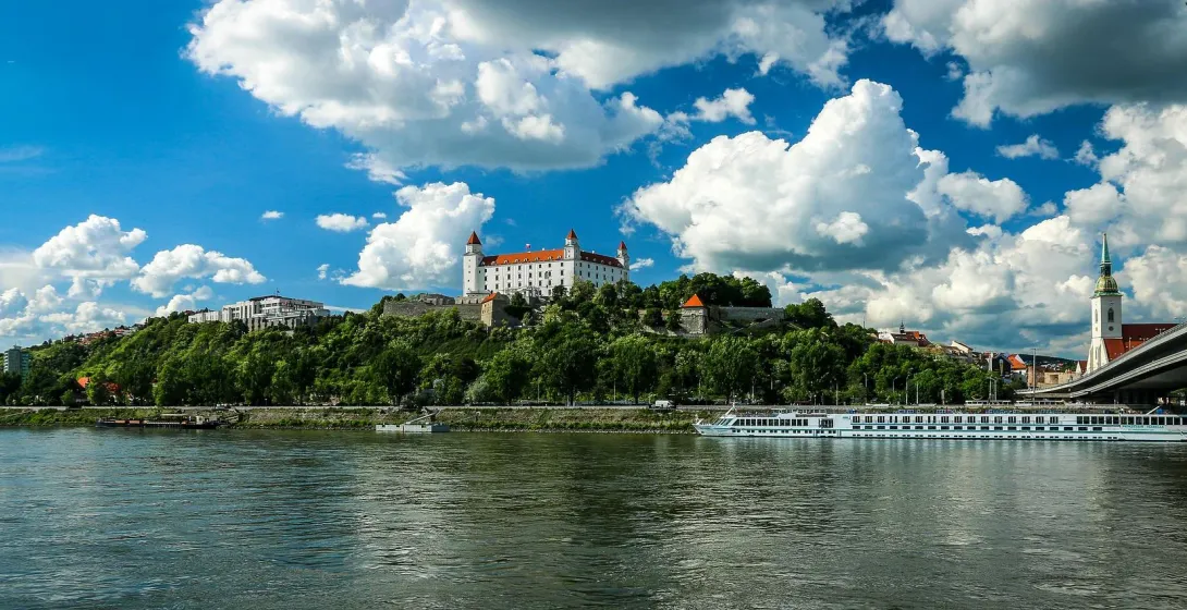 Bratislava Castle on a green hill overlooking the Danube River, with a cruise ship, bridge, and St. Martin's Cathedral spire.