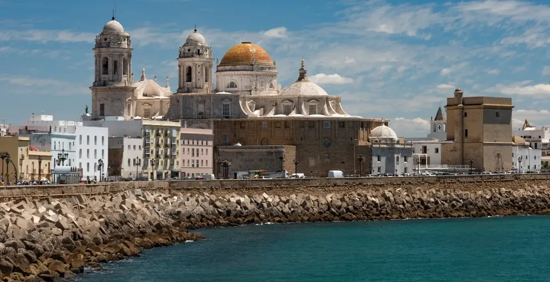 View of Cadiz Cathedral and city skyline from the sea, with a rocky breakwater in the foreground under a blue sky.