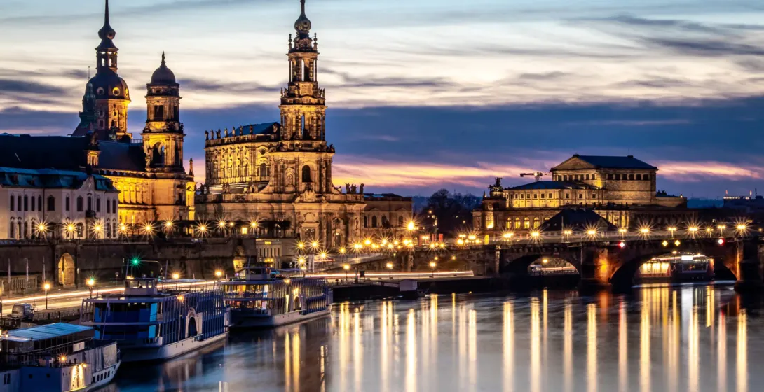 Dresden city skyline at dusk with illuminated historic buildings along the Elbe River, boats docked, and reflections on the w