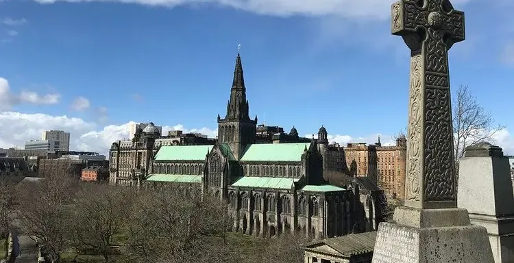 Historic Glasgow Cathedral with green roof and spire, viewed from the Necropolis, showing an ornate Celtic cross in the foreg