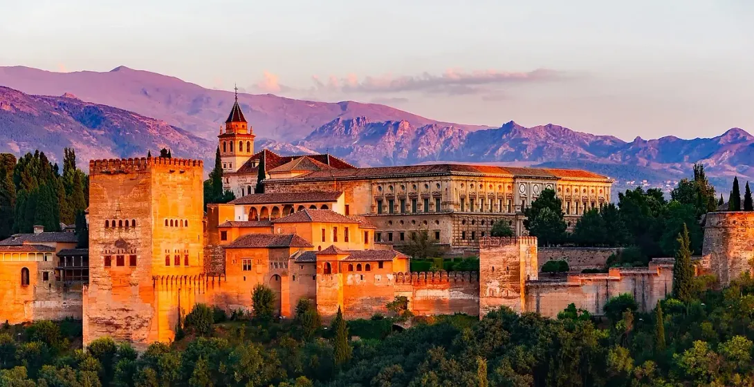 Alhambra palace complex with fortified walls and towers at sunset, mountains in the background, surrounded by green trees.
