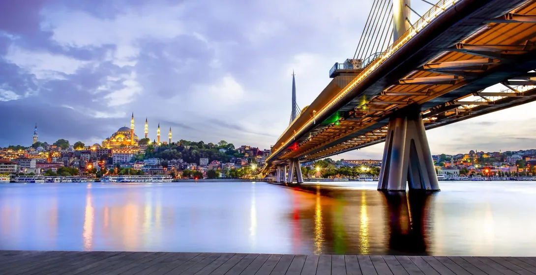 Illuminated Golden Horn Metro Bridge over water at dusk, with the Süleymaniye Mosque and Istanbul cityscape in the background