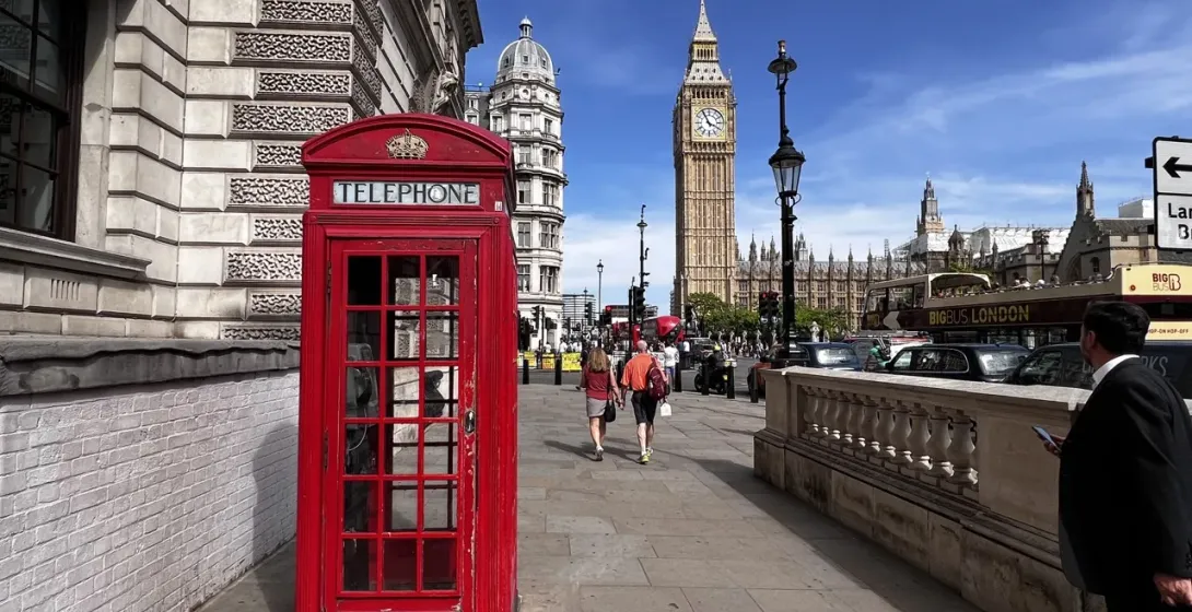A classic red telephone booth on a London sidewalk with Big Ben and the Houses of Parliament in the background under a blue s