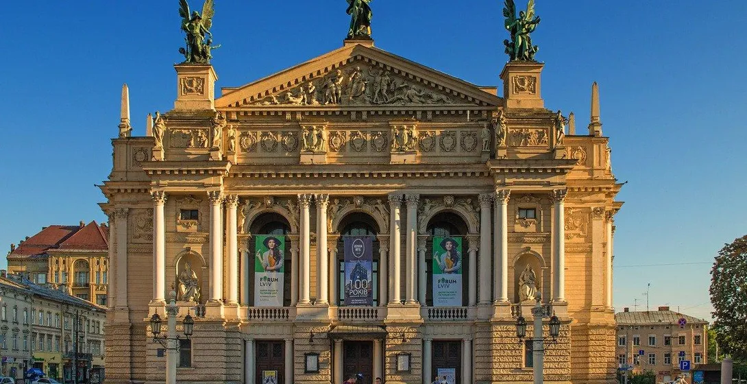 Facade of the Lviv Opera House with columns, sculptures on the roof, and banners. Clear blue sky above. People walk in the fo