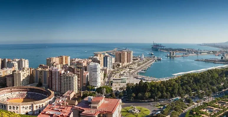 Panoramic view of Malaga cityscape with La Malagueta bullring, the port, and the Mediterranean Sea under a blue sky.