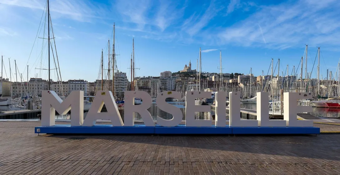 Large MARSEILLE letters on a blue base at the Vieux-Port, with sailboats and buildings. Notre-Dame de la Garde visible on the