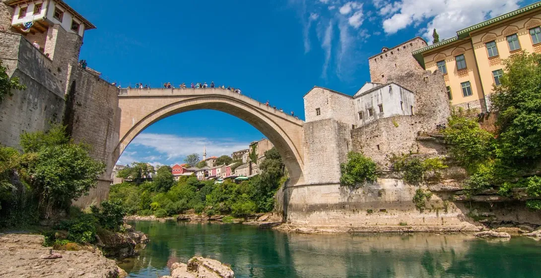 Historic Stari Most bridge in Mostar, Bosnia and Herzegovina, spanning a green river with ancient stone buildings and lush ba