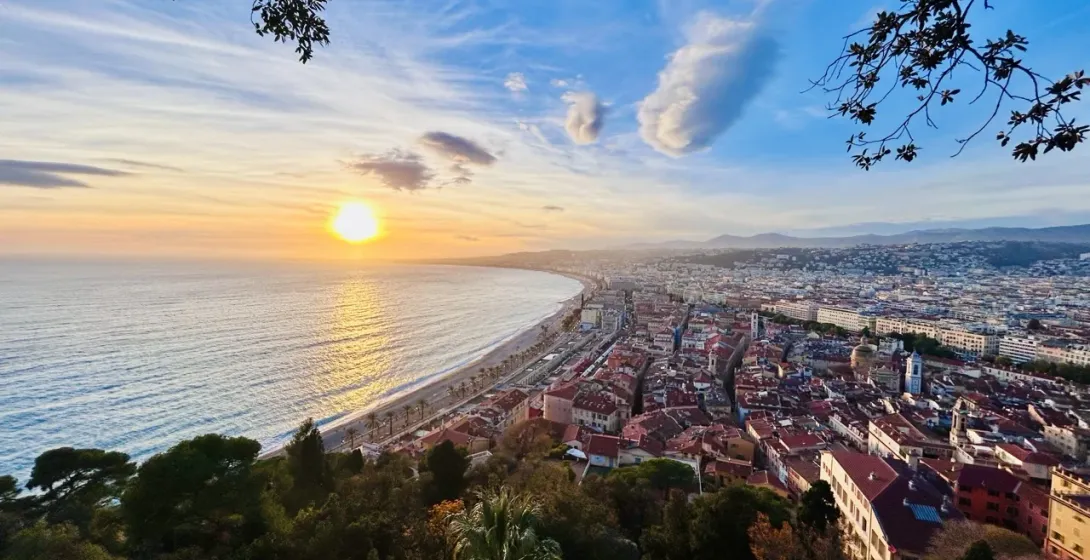 Panoramic view of Nice coastline at sunset, showing the sun reflecting on the sea, a long beach, promenade, and the city buil