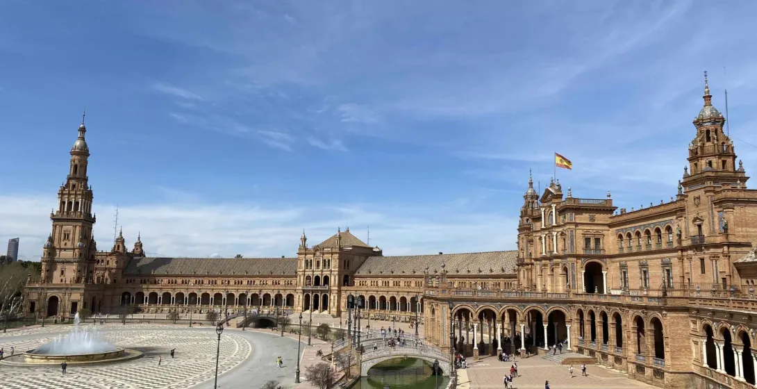 Large ornate building with arcades, a central canal with bridges, patterned plaza, and fountain under a blue sky in Seville.