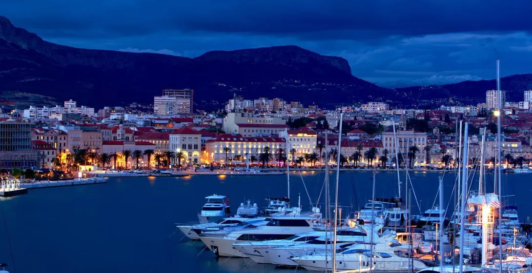 Harbor at twilight with numerous yachts and boats, illuminated waterfront buildings, palm trees, and mountains under a dark b