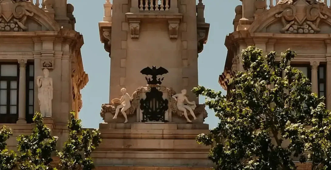 Ornate clock tower of Valencia City Hall with a large clock face, statues, flags, and green trees in foreground under a clear
