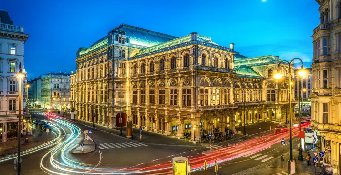 Illuminated Vienna State Opera building at twilight, with light trails from cars on a street crossing and a crescent moon in