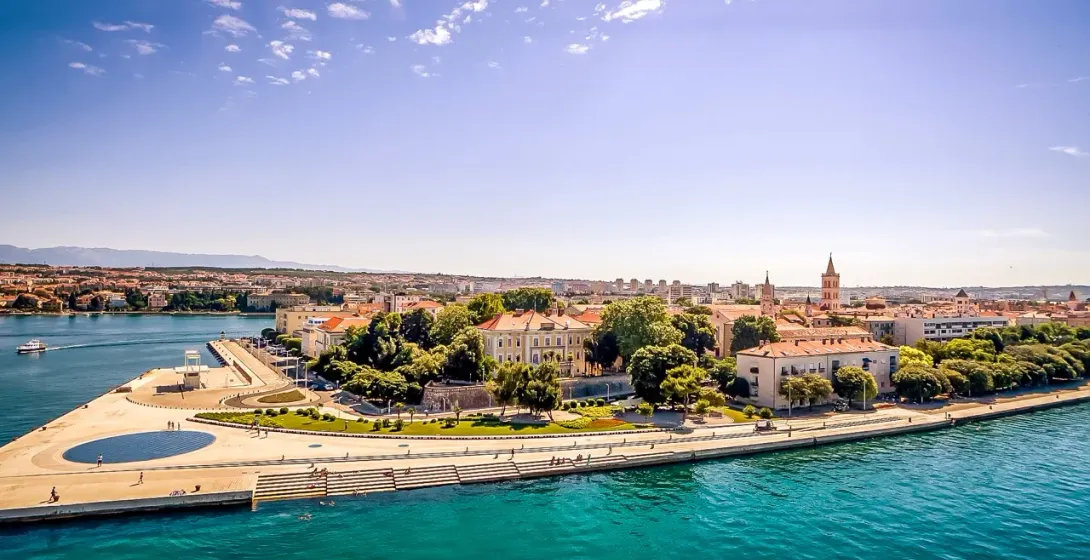 Aerial view of Zadar waterfront with the Sea Organ, old town buildings, clear blue sea, and distant mountains under a sunny s