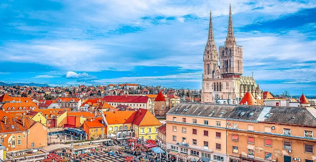 Aerial view of Zagreb with the Cathedral featuring two spires, partially scaffolded, and a bustling outdoor market with red u