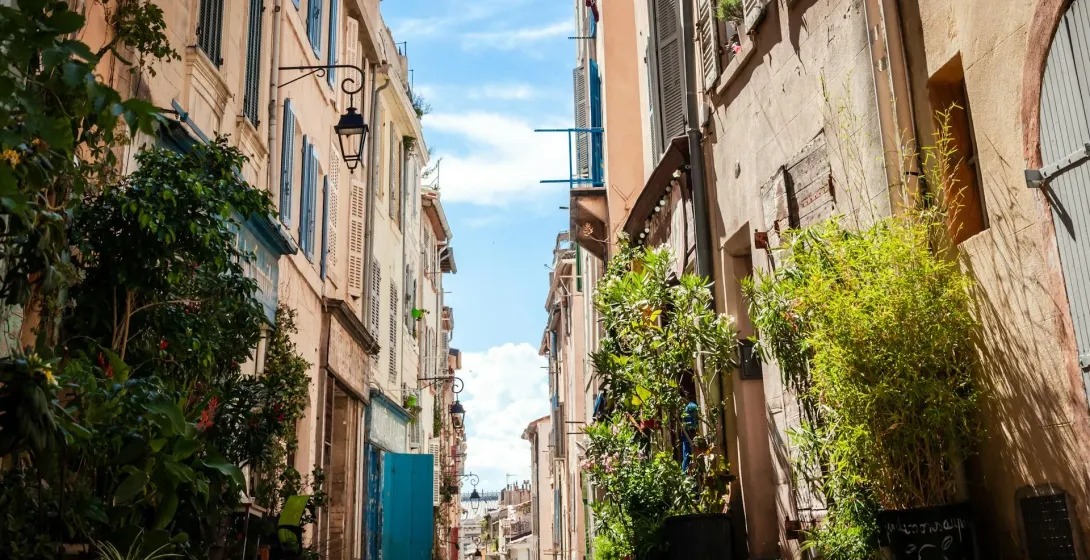 Narrow paved alleyway in Marseille lined with tall old buildings, closed shutters, and green potted plants, under a blue sky.