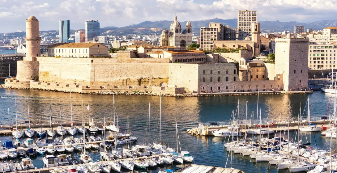Panoramic view of Marseille's Old Port with Fort Saint-Jean, a lighthouse tower, and numerous boats. City skyline with the ca