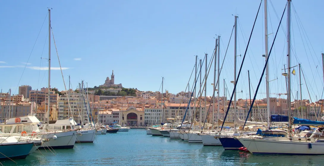Sailboats and yachts docked in a blue harbor, with a city skyline and Notre-Dame de la Garde basilica on a hill in the backgr