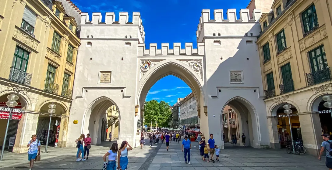 The Karlstor archway in Munich, a white stone gate with two crenellated towers and three passages, with people walking throug