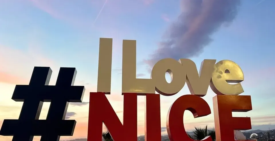 Large 3D 'I Love #NICE' sign with a dark blue hashtag, gold 'I Love', and red 'NICE' against a pastel sunset sky over water.