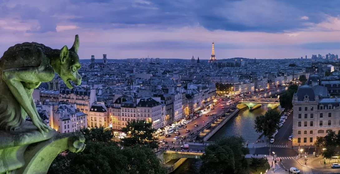Stone gargoyle on a building overlooking the Paris cityscape at dusk, with the illuminated Eiffel Tower and city lights.