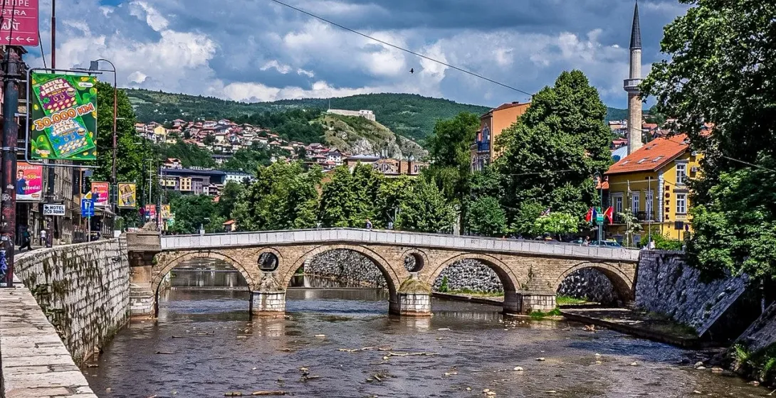Historic stone arch bridge over a river in Sarajevo, with a minaret, buildings, and green hills under a cloudy sky.
