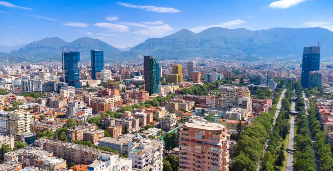 Aerial view of Tirana city with modern high-rise buildings, residential areas, a tree-lined boulevard, and mountains.
