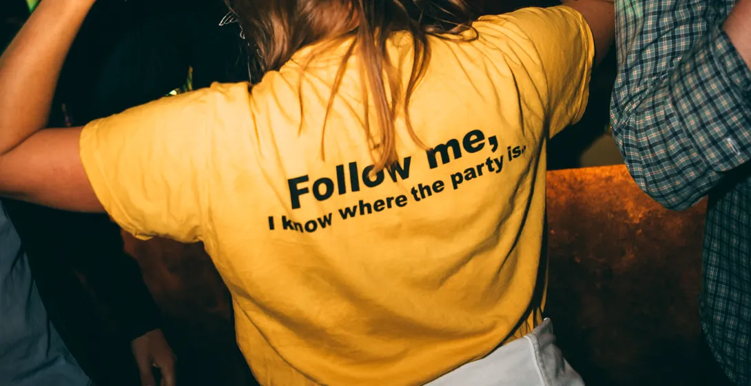 People dancing in a dimly lit bar, one person in a yellow t-shirt with 'Follow me, I know where the party is' on the back.